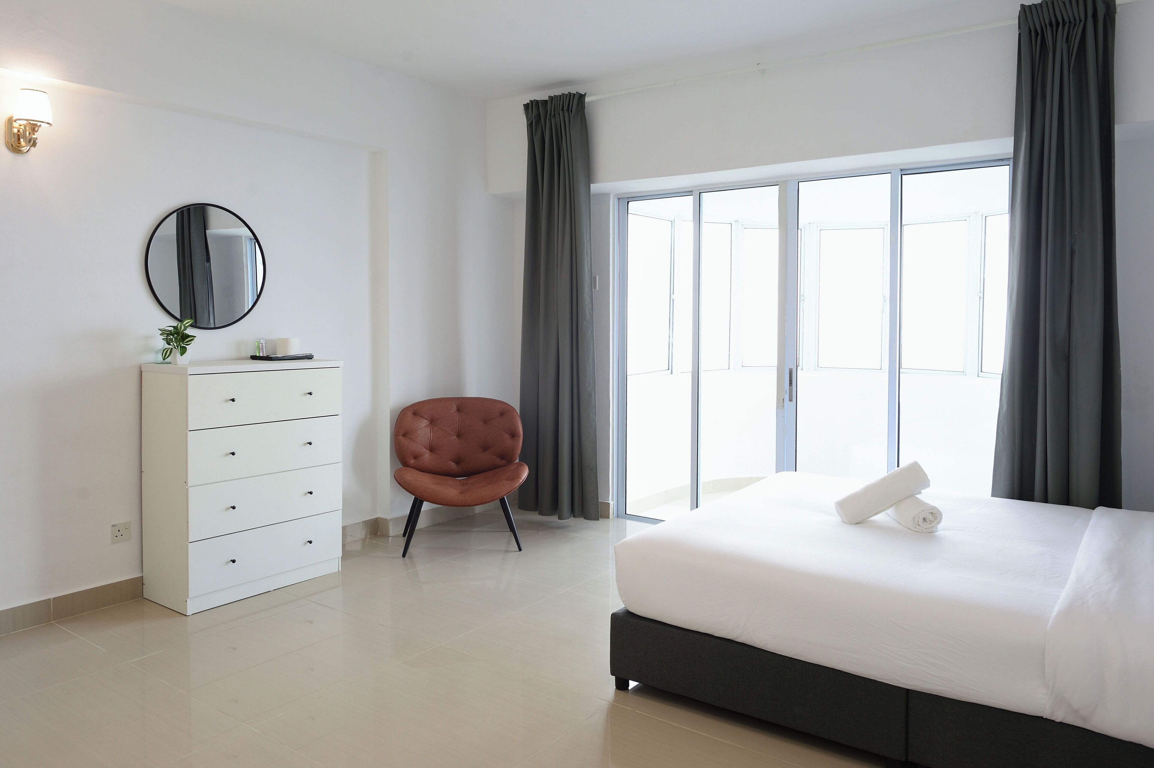 Un dormitorio moderno con una cama grande, un tocador, una silla y un ventanal corredizo que da a un balcón.