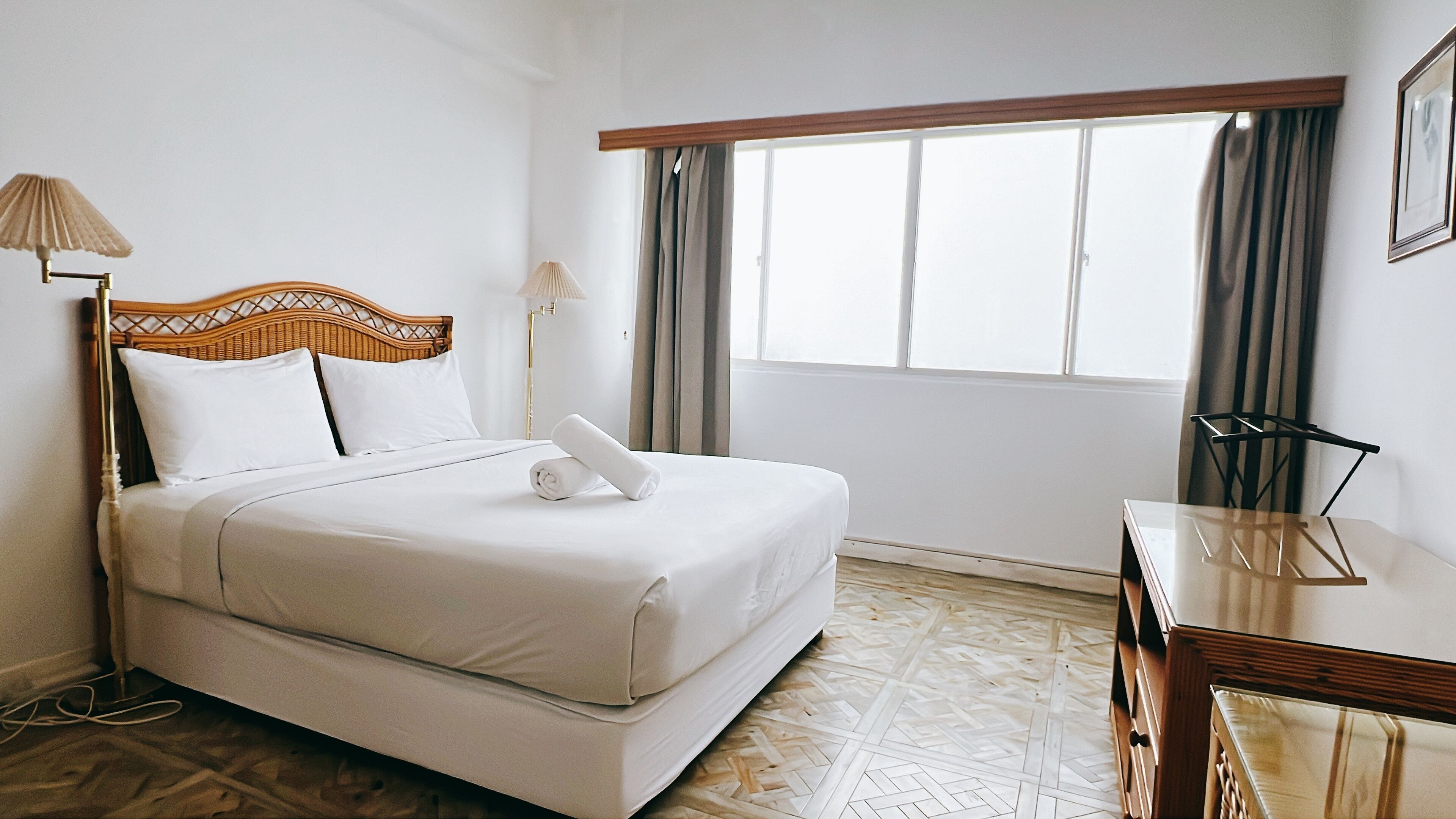 A neatly made bed with white linens and a rolled towel, a wooden headboard, a bedside table with a lamp, and a window with curtains.