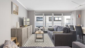 A modern living room with a grey sofa, a wooden coffee table, and a TV on a wooden cabinet. Large windows provide a view of the city. - Pleasant apartment on the 2nd floor (Blankenberge)