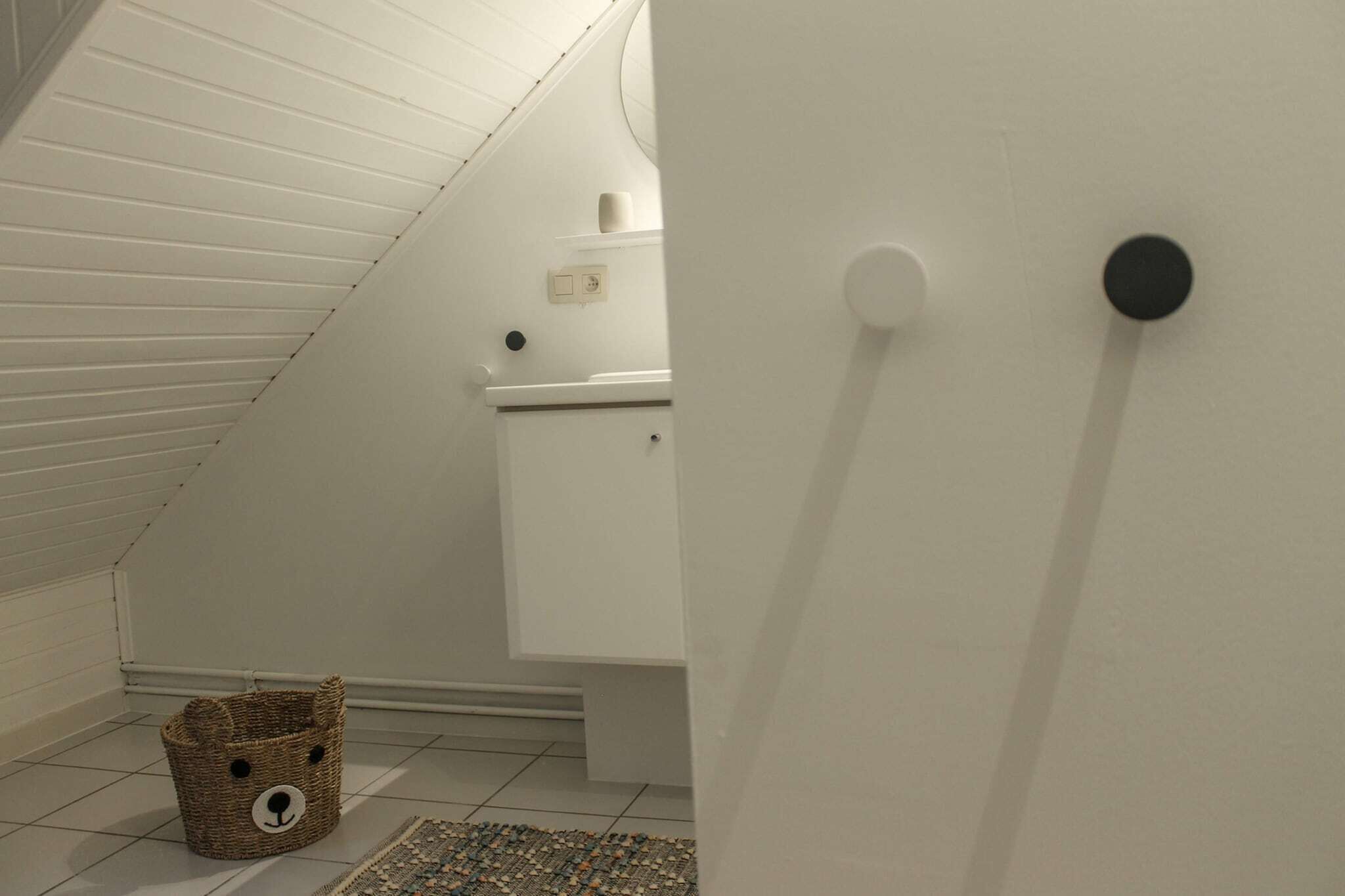 A white interior with a sloped ceiling, a small wicker basket with a face design, a white cabinet, and a round wall-mounted object.
