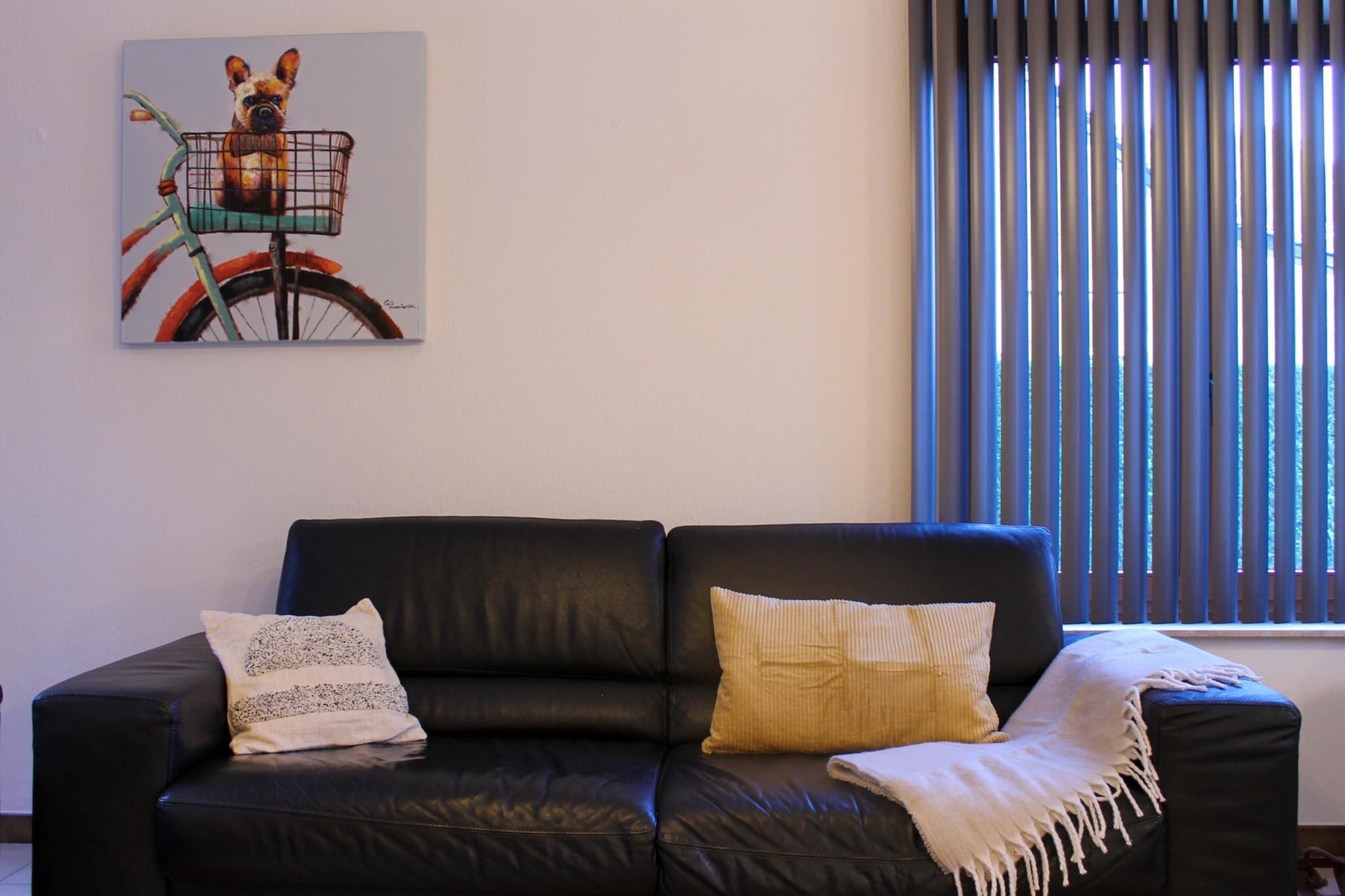 A black leather sofa with three cushions, a framed artwork of a dog in a basket on a bicycle, and a window with vertical blinds.