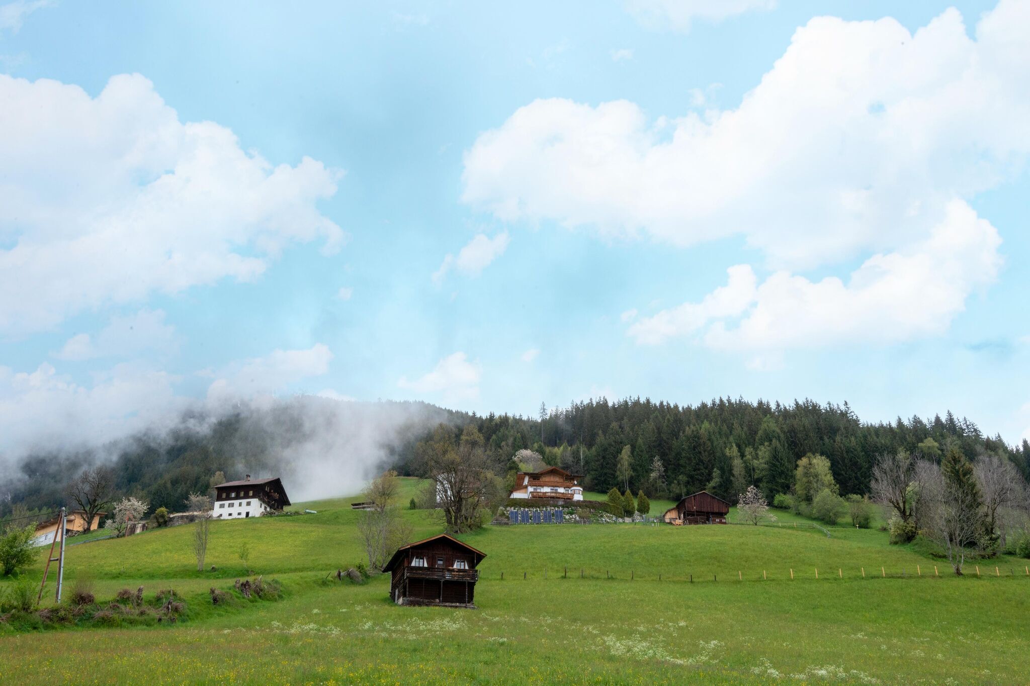 A rural landscape with several buildings, including a large white house and smaller structures, surrounded by green fields and a forested hill.