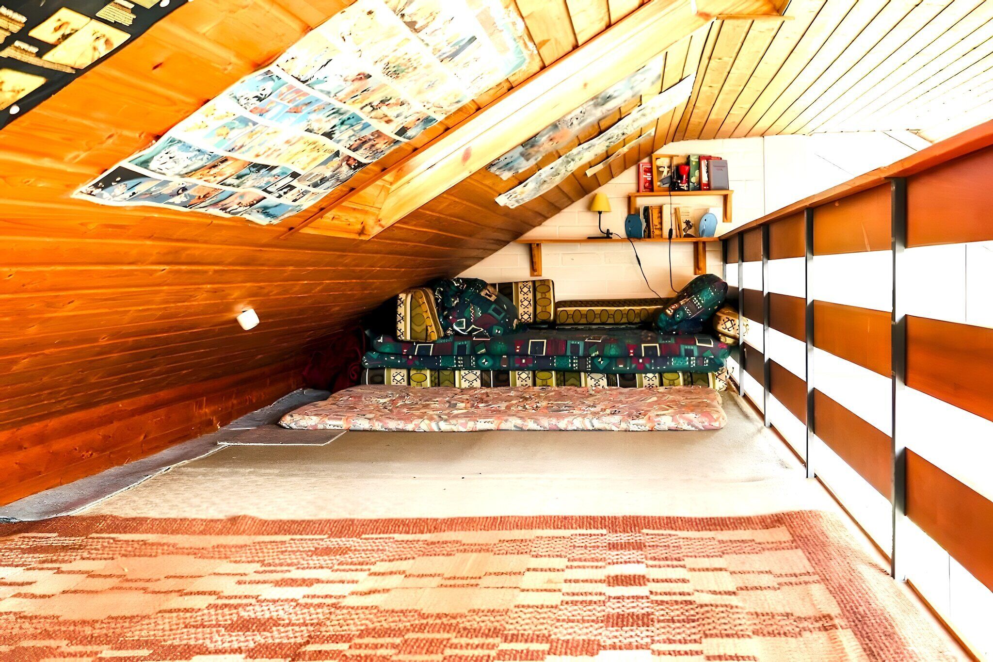 A cozy attic bedroom with a sloped ceiling, a patterned rug, a colorful sofa, and a bookshelf.