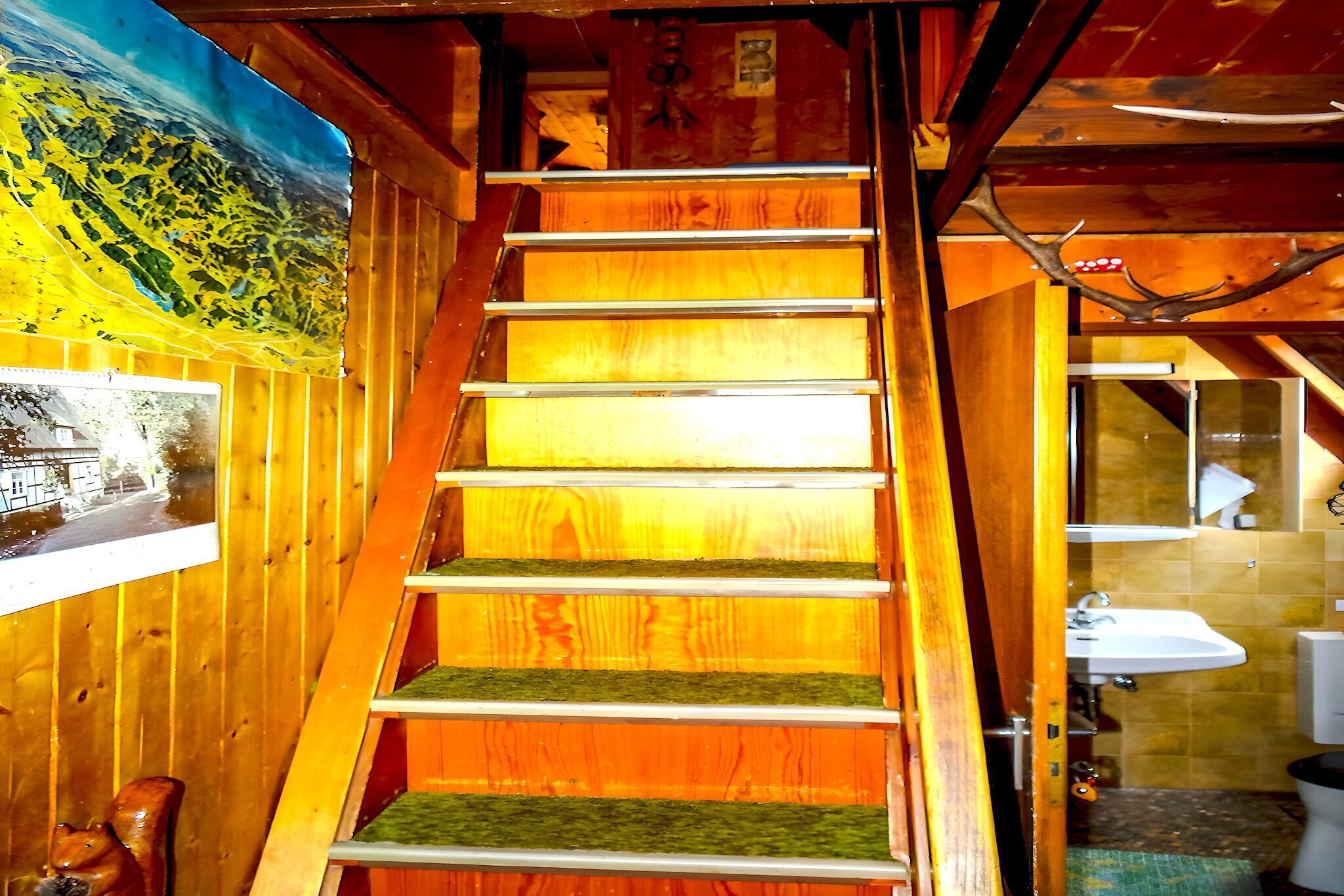 Un escalier en bois avec un palier recouvert d’une moquette verte, un tableau représentant un paysage et une salle de bain où l’on peut voir un lavabo et des toilettes.