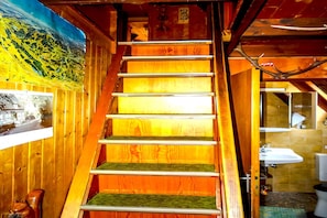 A wooden staircase with a green carpeted landing, a painting of a landscape, and a bathroom with a sink and toilet visible.