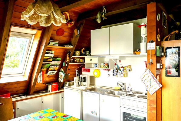 Une cuisine de taille réduite, avec des meubles blancs, une cuisinière et un évier. Une fenêtre laisse entrer la lumière naturelle et une porte donne accès à une autre pièce.