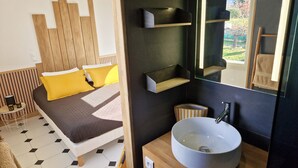 Een moderne slaapkamer met een houten hoofdbord, een bed met gele en bruine beddengoed, een nachtkastje met een lamp, en een grote spiegel boven een badkamermeubel.