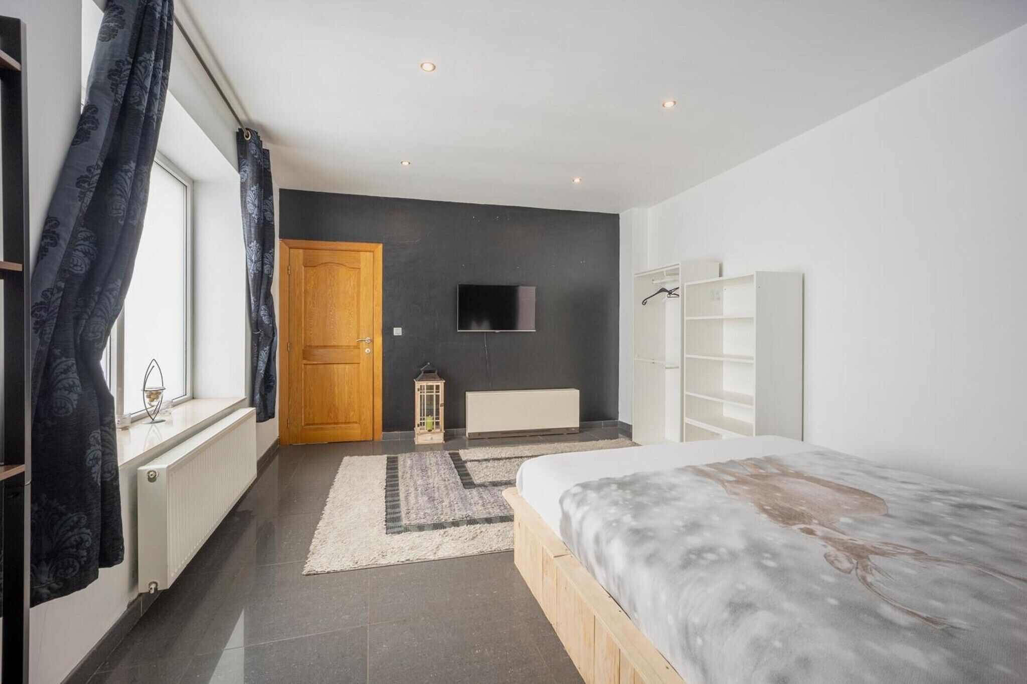 A bedroom with a bed, a TV, a radiator, a wooden door, and a window with curtains.