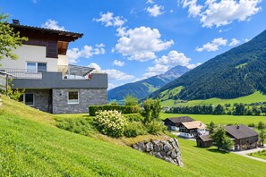 A modern two-story house with a stone facade, a balcony, and a view of a mountainous landscape.