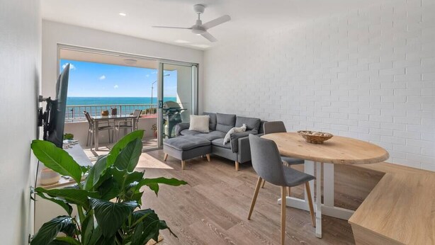 Una sala de estar moderna con un sofá seccional gris, una mesa de comedor redonda de madera y un balcón con vista al mar.