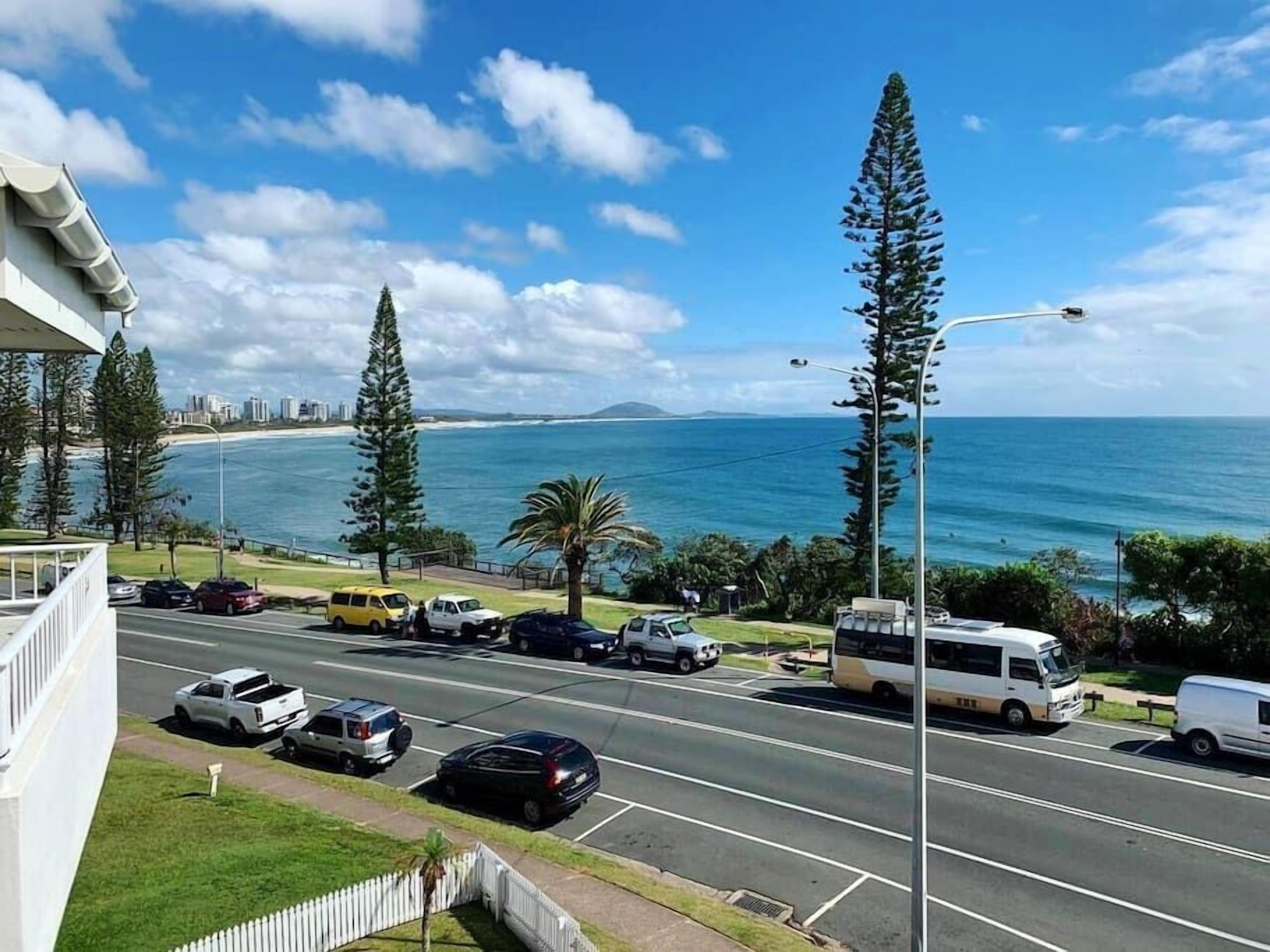 A coastal road with parked vehicles, a bus, and tall trees.