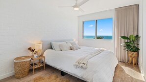 Un dormitorio con una cama, un ventilador de techo, una mesita de noche con lámpara, una ventana con vista al mar y una planta en maceta.
