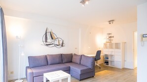 A modern living room with a grey sectional sofa, a white coffee table, and a floor lamp. There is a white shelving unit with decorative items and a small desk with a chair. - Duplex apartment near the sea (Middelkerke)