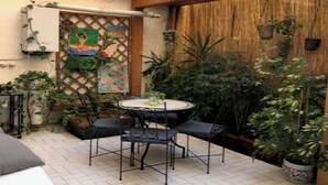 Um pátio com mesa redonda e cadeiras, rodeado por plantas em vasos e uma treliça de madeira.