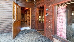 A wooden cabin with a wooden porch, a door leading to a room, and a window with a checkered curtain.