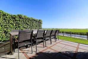 Un patio al aire libre con mesa y sillas, un seto y vista a un cuerpo de agua.