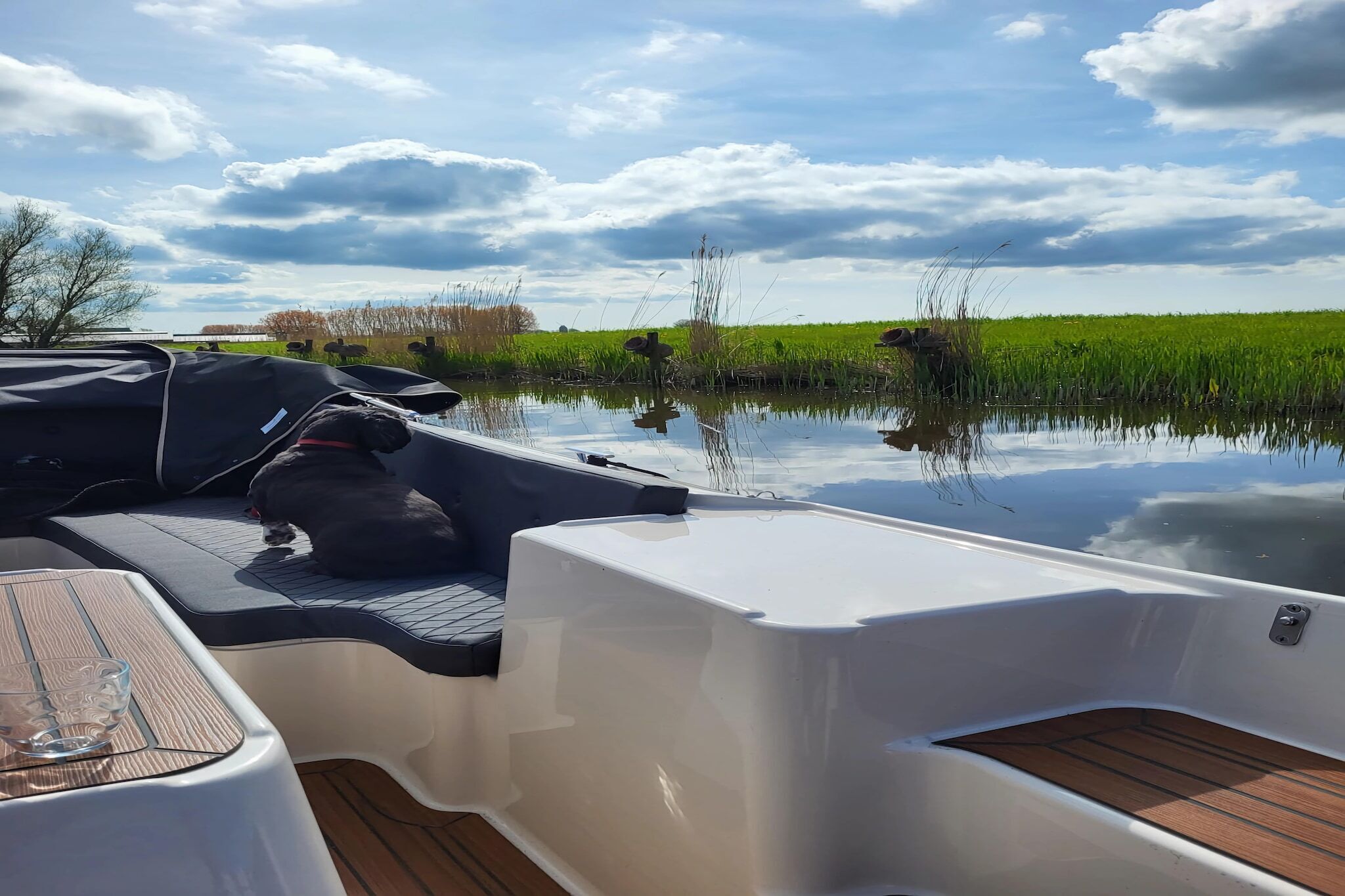 Le pont d’un bateau, avec une table et un espace abrité, entouré d’une eau calme et d’un champ verdoyant sous un ciel nuageux.