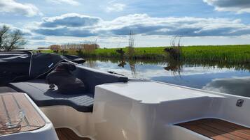 Le pont d’un bateau, avec une table et un espace abrité, entouré d’une eau calme et d’un champ verdoyant sous un ciel nuageux.