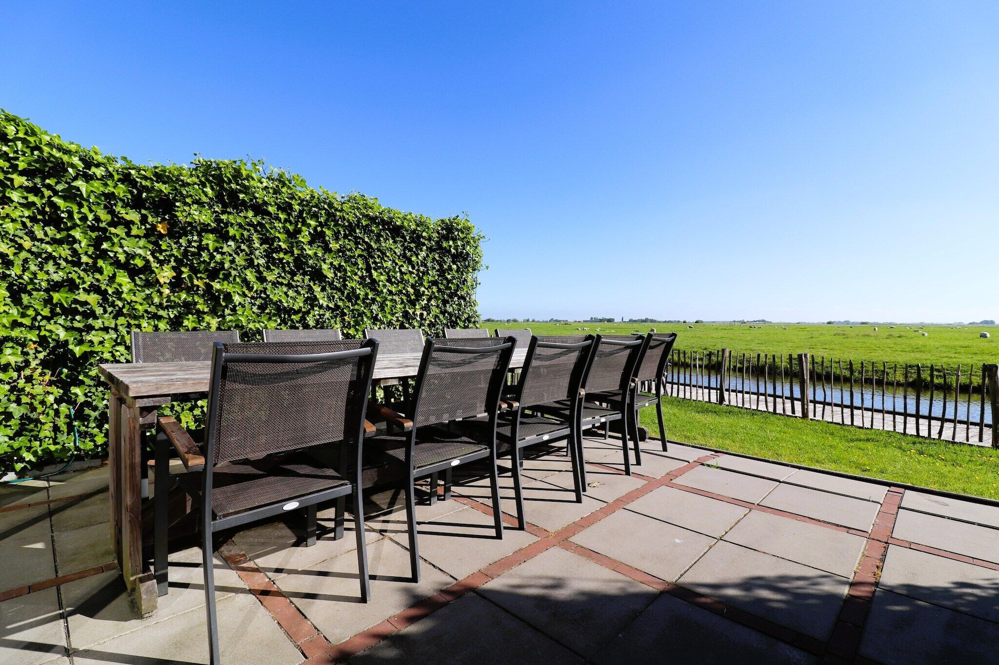 A patio with outdoor seating, a hedge, and a view of a water body.