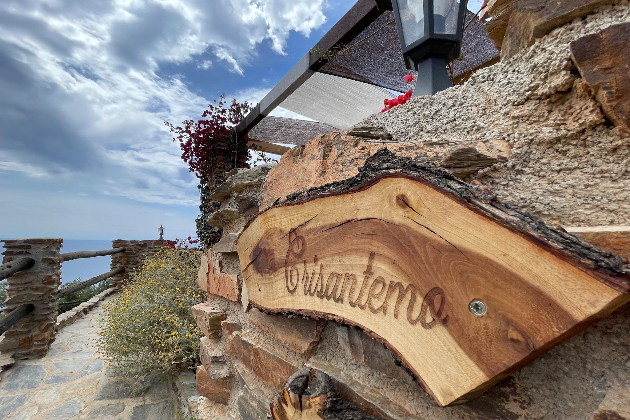 A wooden sign with the name "Crisantino" carved into it, mounted on a stone wall with a view of the sea in the background.