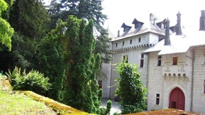Un edificio histórico de piedra con puerta roja, rodeado de abundante vegetación y altos árboles.