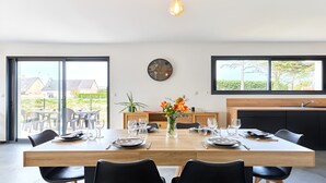 Un espace repas moderne avec une table en bois dressée pour six personnes, un vase de fleurs et une grande fenêtre donnant sur des maisons.
