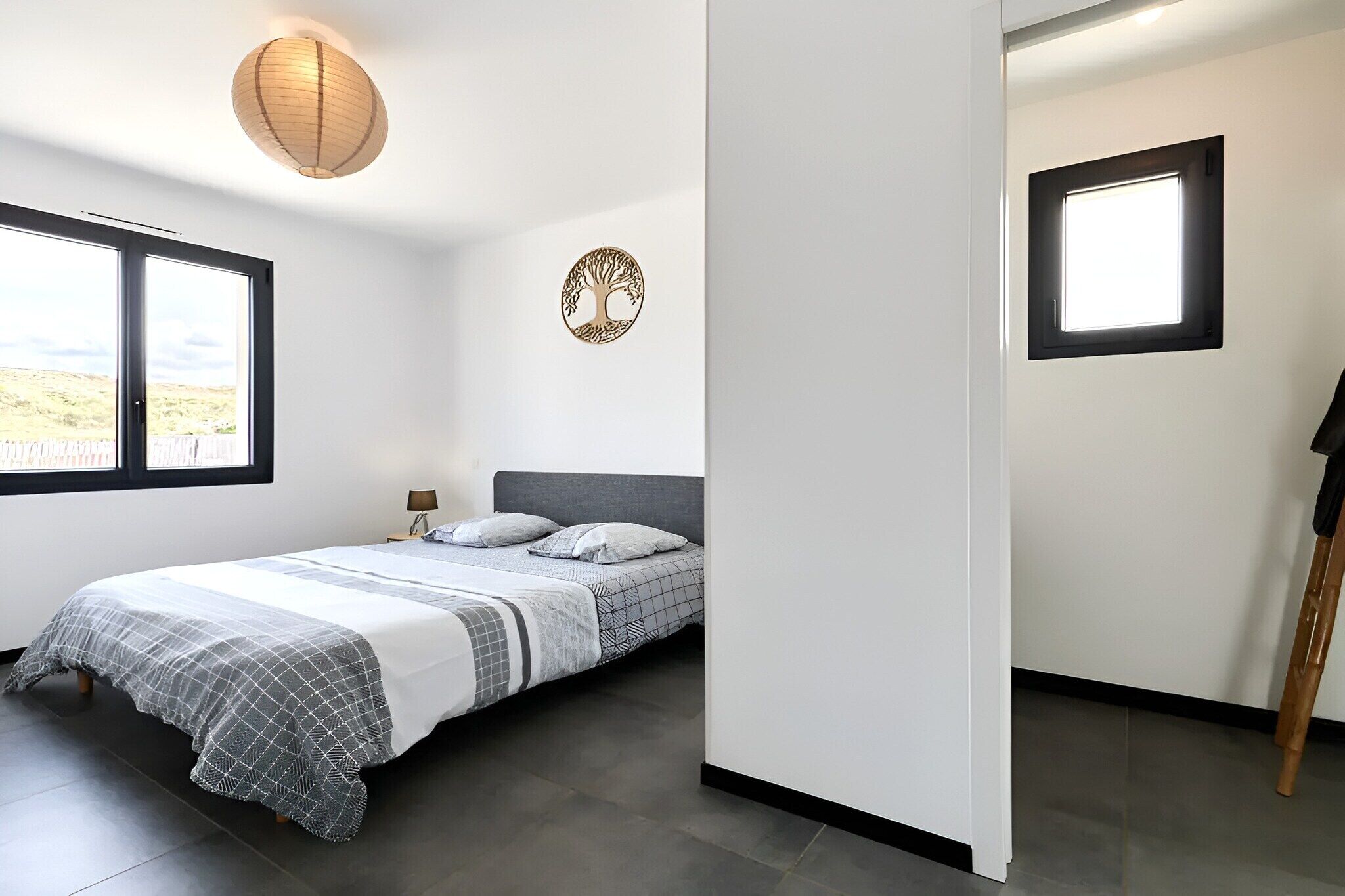A bedroom with a bed, a window, a wall-mounted lamp, and a small window.