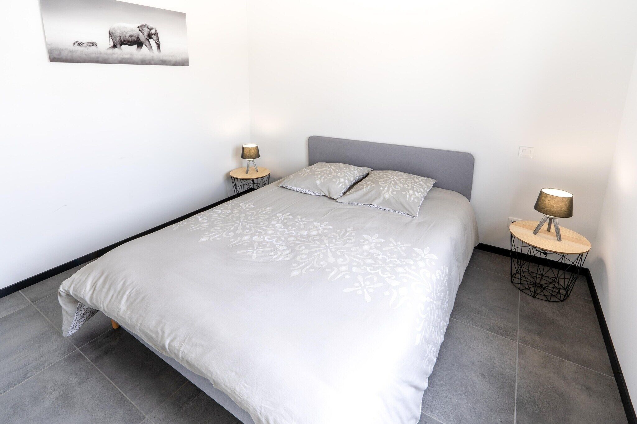 Ein Bett mit grauem Kopfteil, zwei Nachttische mit Lampen sowie ein Wandbild von Elefant und Zebra.