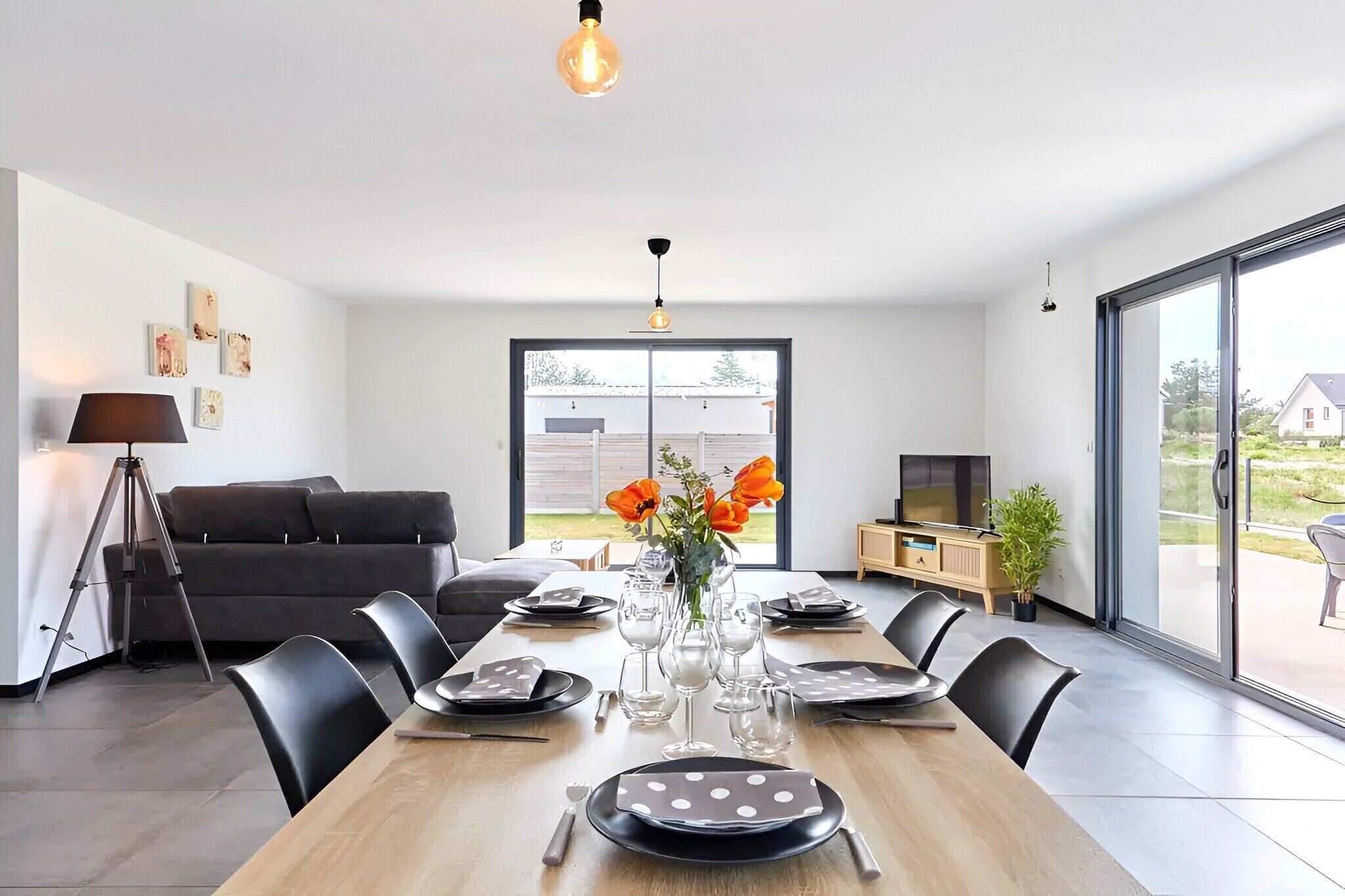 A modern dining area with a wooden table set for six, a sofa, a TV, and a large window.