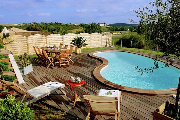 A wooden deck with a swimming pool, lounge chairs, and a table with a red tablecloth.
