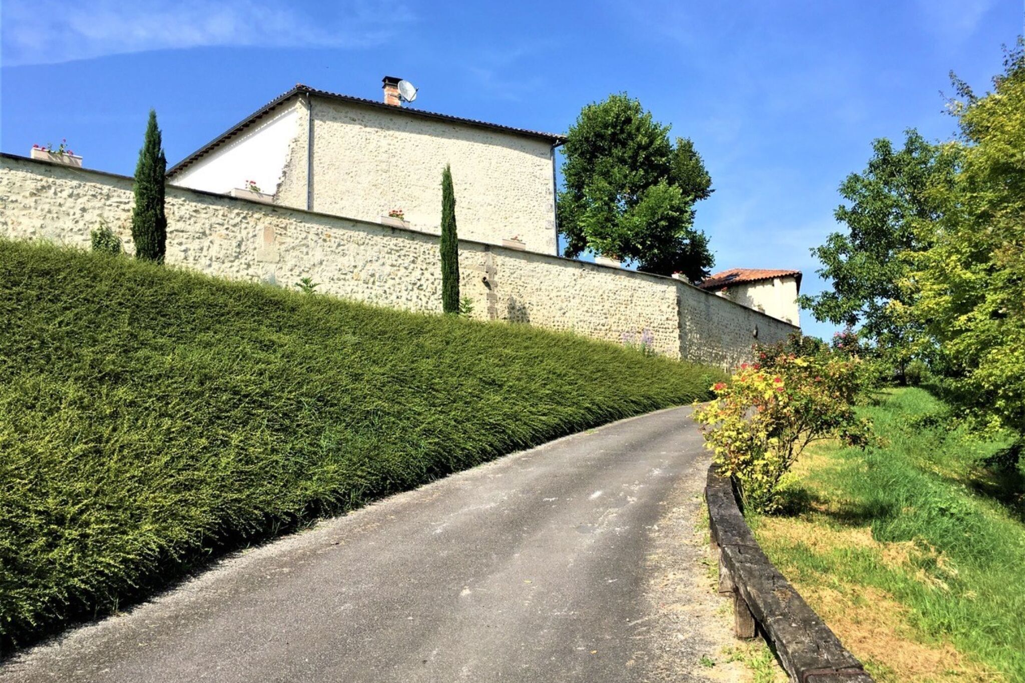Un chemin étroit mène à un bâtiment en pierre surmonté d’un toit de tuiles, entouré de haies et d’arbres bien entretenus.