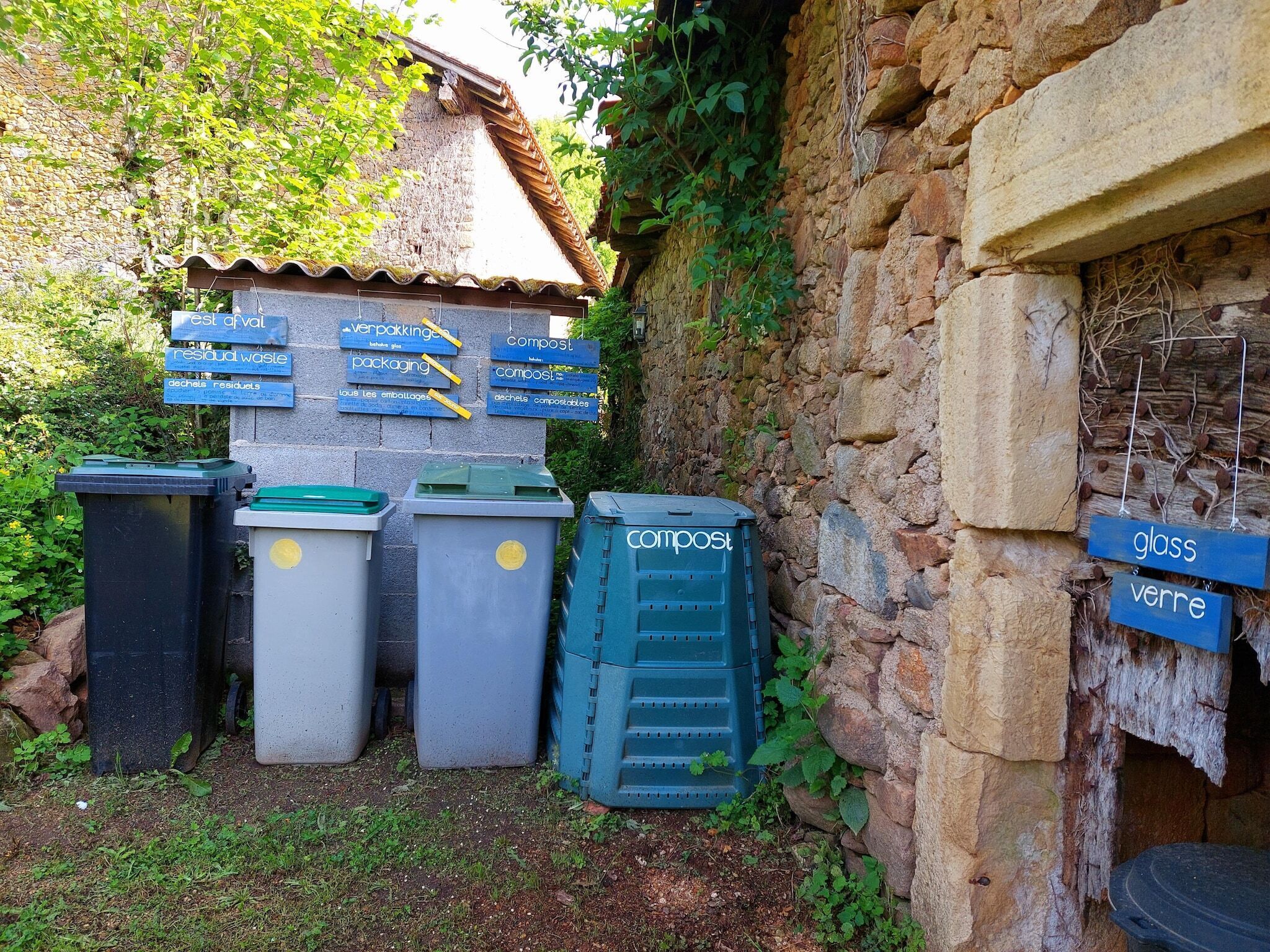 Outdoor recycling bins with labels for glass, plastic, and compost, and a stone wall with a rustic appearance.