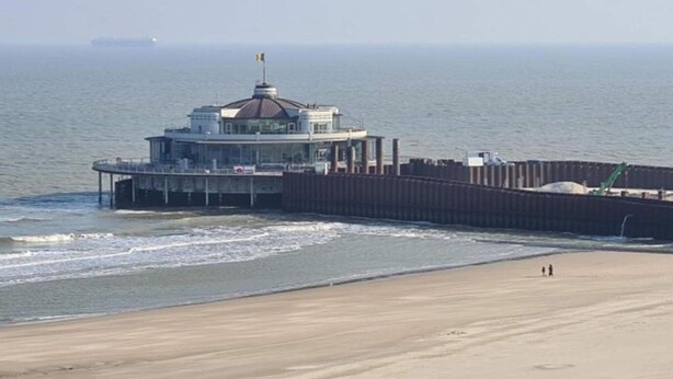 Um edifício costeiro com cúpula, um cais que se estende para a água e uma praia arenosa.