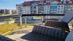 Uma varanda com cadeiras de vime, guarda-corpo de vidro e vista para edifícios residenciais.
