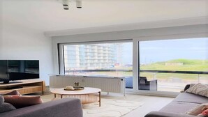Uma sala de estar moderna com uma janela ampla, uma televisão de tela plana, uma mesa de centro redonda e um sofá.
