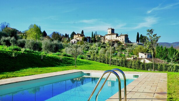Une piscine à l’eau d’un bleu limpide, entourée d’une terrasse dallée. Au fond, on aper çoit une colline parsemée de bâtiments, dont une structure imposante en forme de tour.