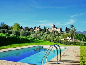 Ein Schwimmbecken mit klarer, blauer Wasseroberfläche, umgeben von einer gepflasterten Terrasse. Im Hintergrund erstreckt sich eine Hügelkuppe mit mehreren Gebäuden, darunter ein markantes, turmartiges Bauwerk.