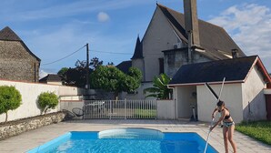 Una piscina con alguien limpiándola, rodeada por un muro de piedra, un árbol pequeño y un edificio con techo a dos aguas.