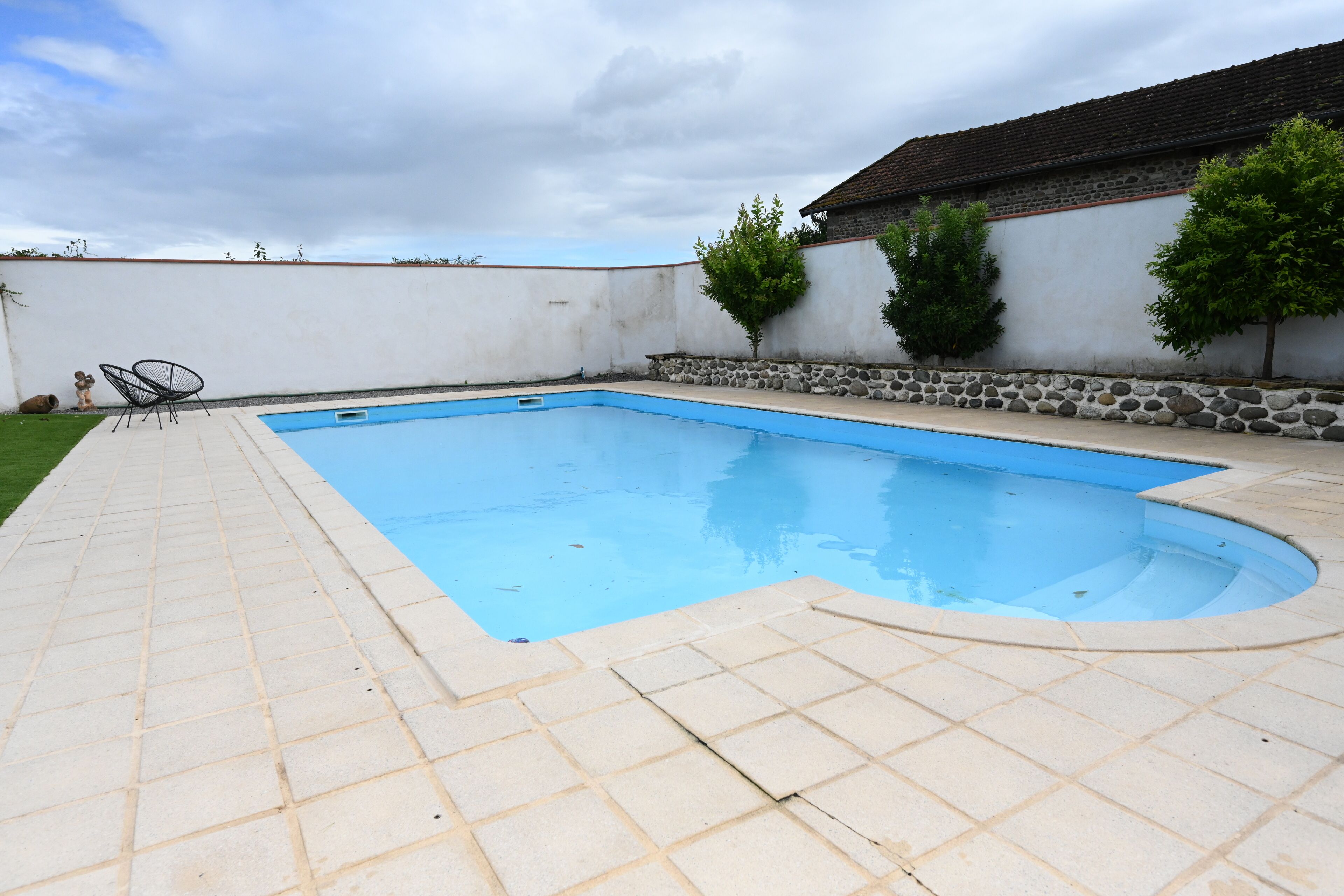 A rectangular swimming pool with a tiled deck, surrounded by a low stone wall and a white building with a tiled roof.