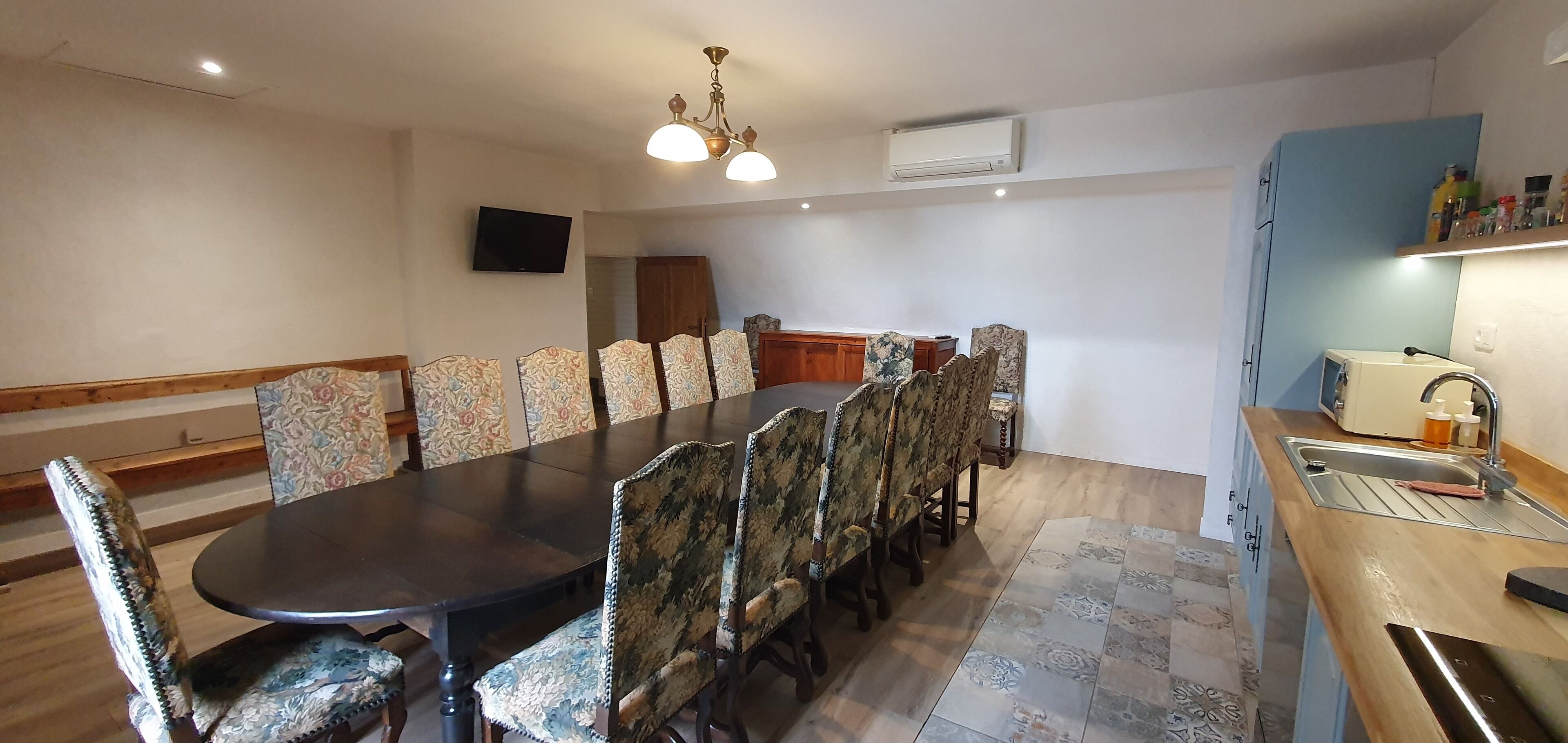 A dining area with a long table and patterned chairs, a kitchen with a sink and countertop, and a ceiling light fixture.