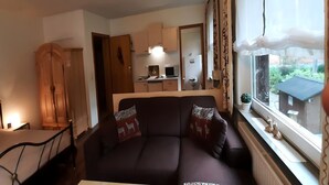 A living room with a sofa, a wooden wardrobe, a kitchen area with cabinets and appliances, and a window with curtains. - Ferienwohnung in Wildemann (Wildemann)