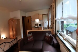 Ein Wohnzimmer mit einer Couch, einem Holzschrank, einer Küchenzeile mit Schränken und Geräten sowie einem Fenster mit Vorhängen.