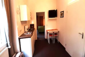 Een smalle gang met een kitchenette, een eethoek met twee stoelen, een aan de muur bevestigde televisie en een deur naar een andere kamer.