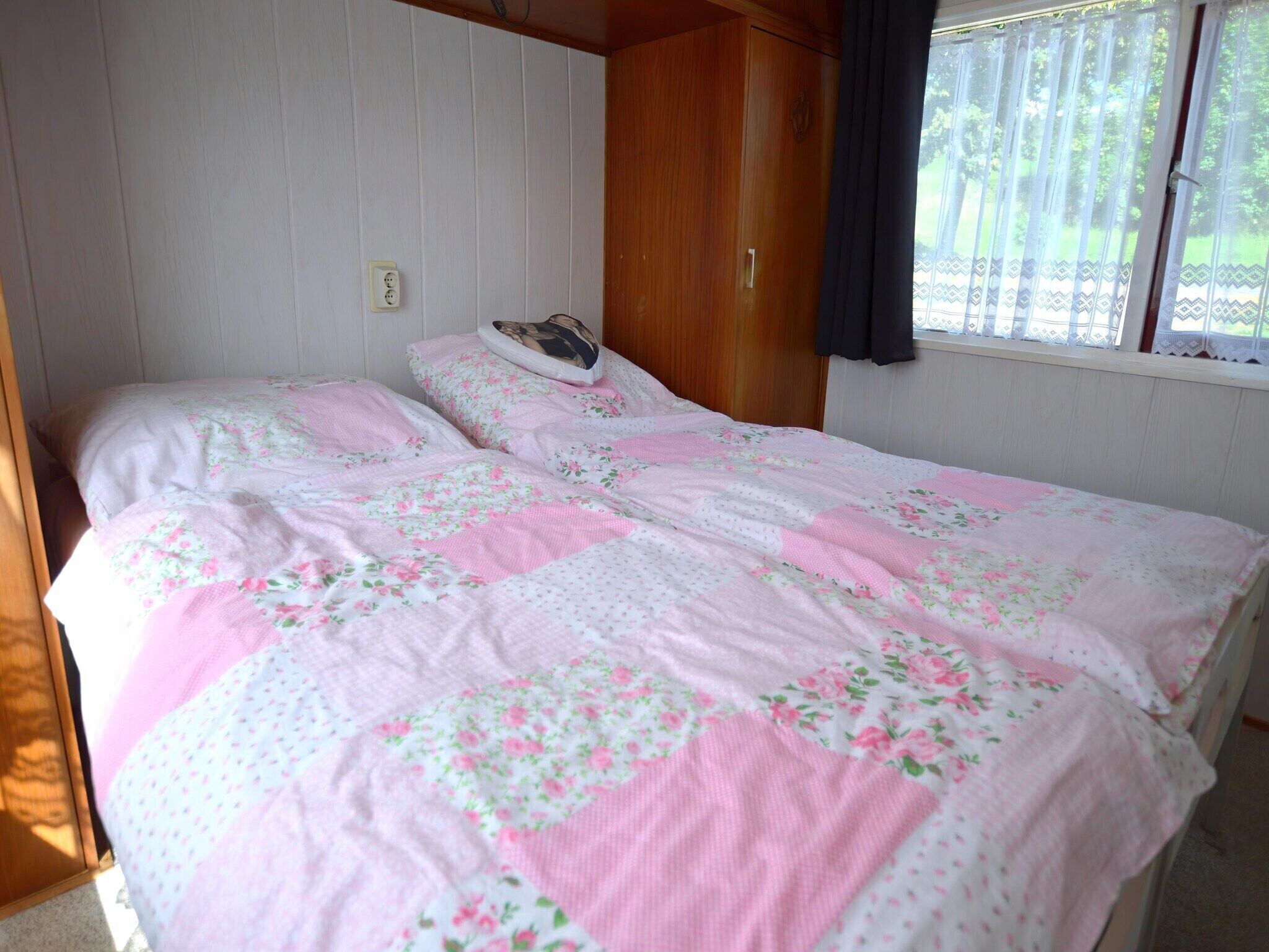 A bedroom with a bed covered in floral bedding, a wooden wardrobe, and a window with curtains.
