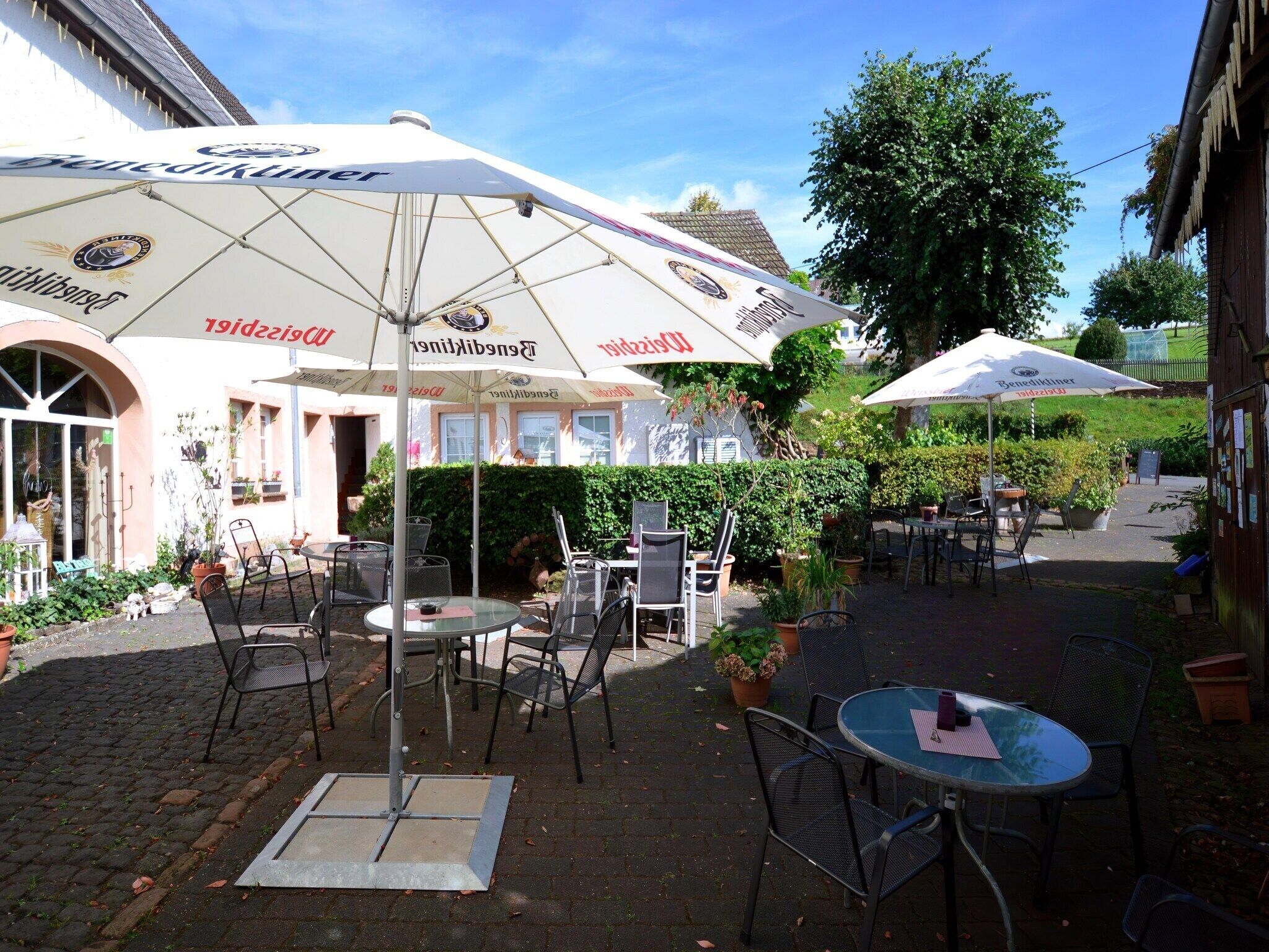 An outdoor café with tables and chairs under umbrellas, surrounded by greenery and a building with signage.