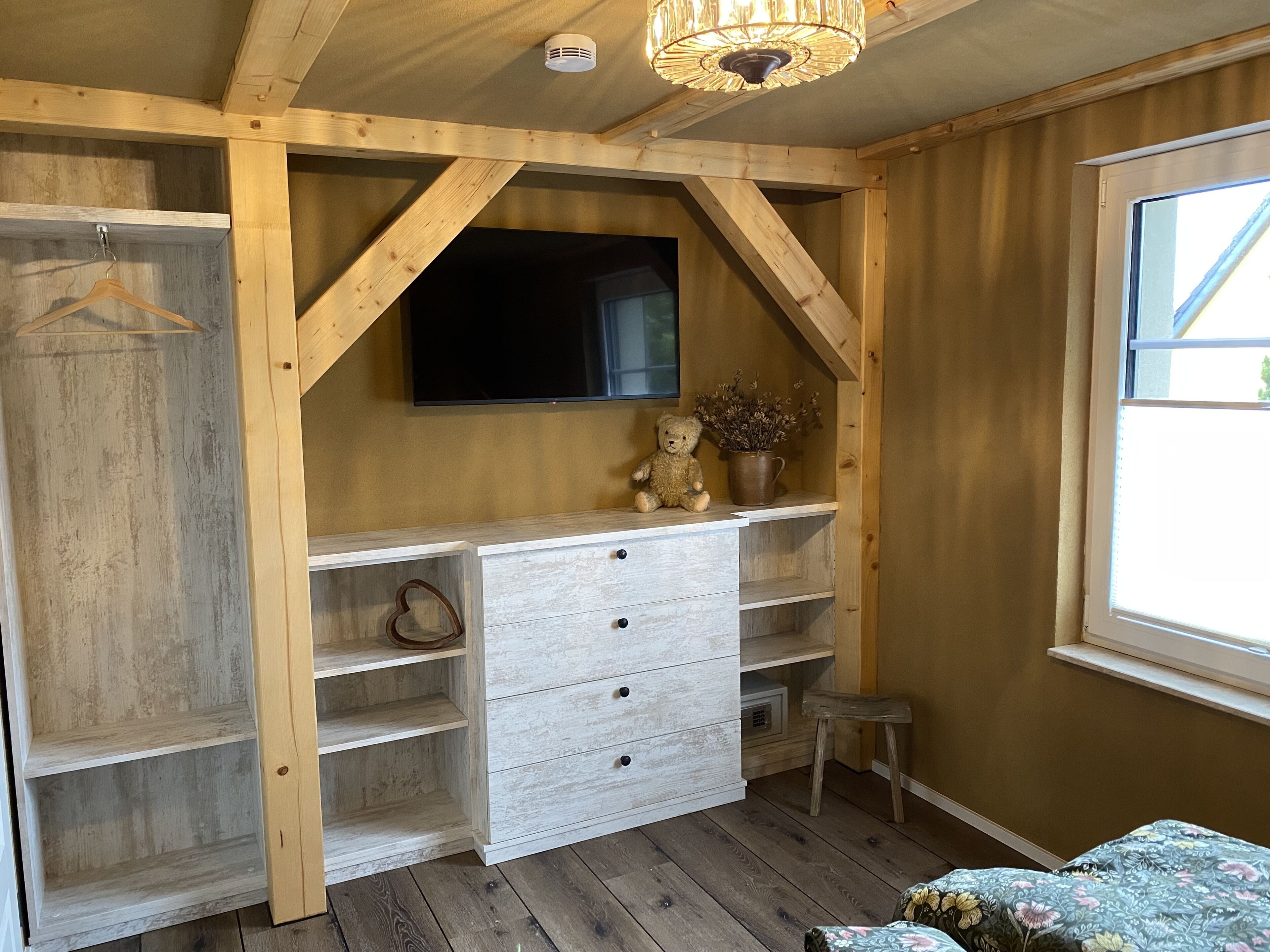 A room with a wooden ceiling, a television mounted on the wall, a dresser with drawers, and a window with a view.