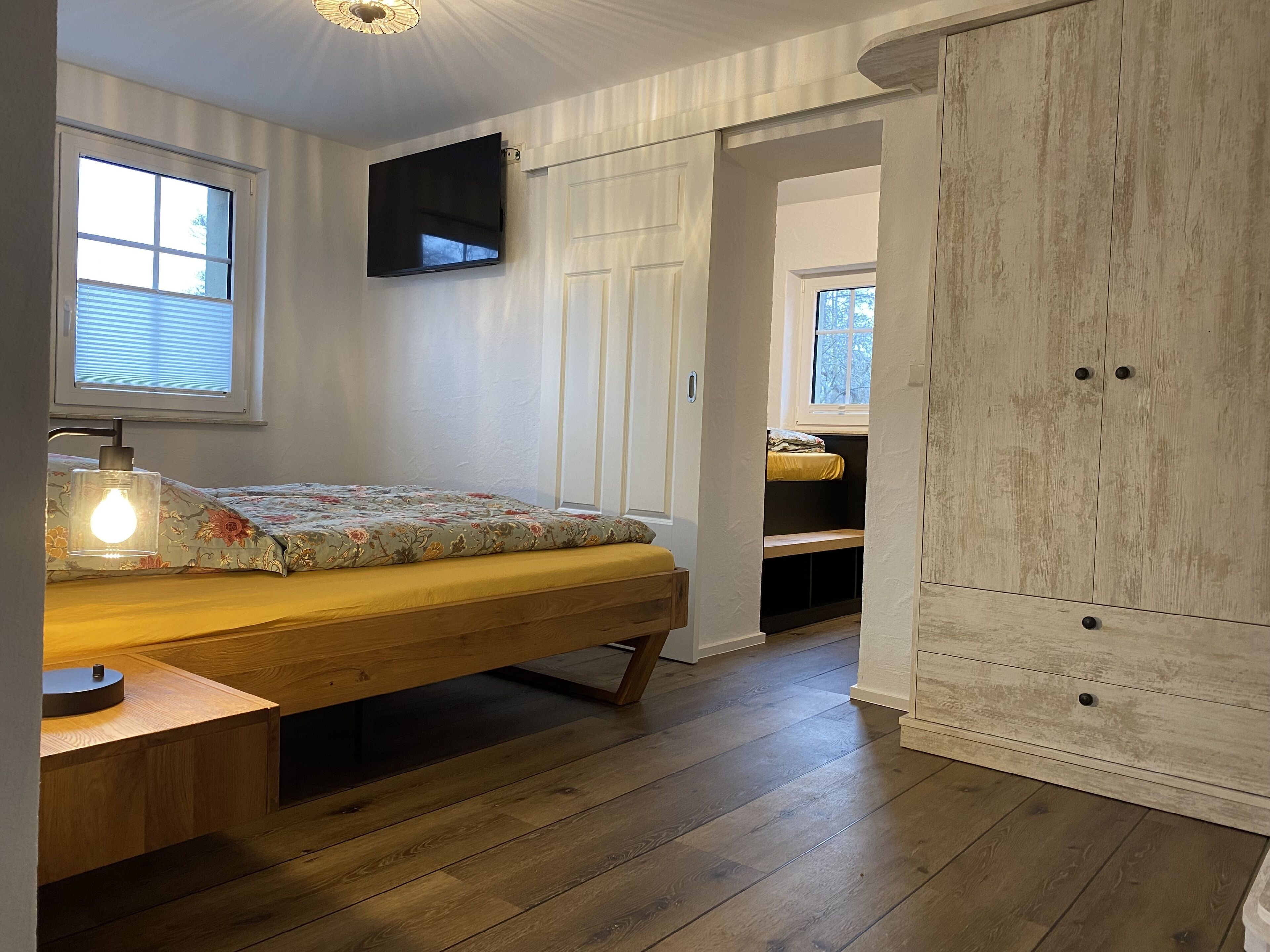 Une chambre à coucher avec un lit en bois, une lampe de chevet, un téléviseur fixé au mur et une armoire.