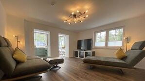 A spacious living room with wooden flooring, two sofas with cushions, a television on a stand, and a ceiling light fixture. - Haus Stenzinger (Burg (Spreewald))