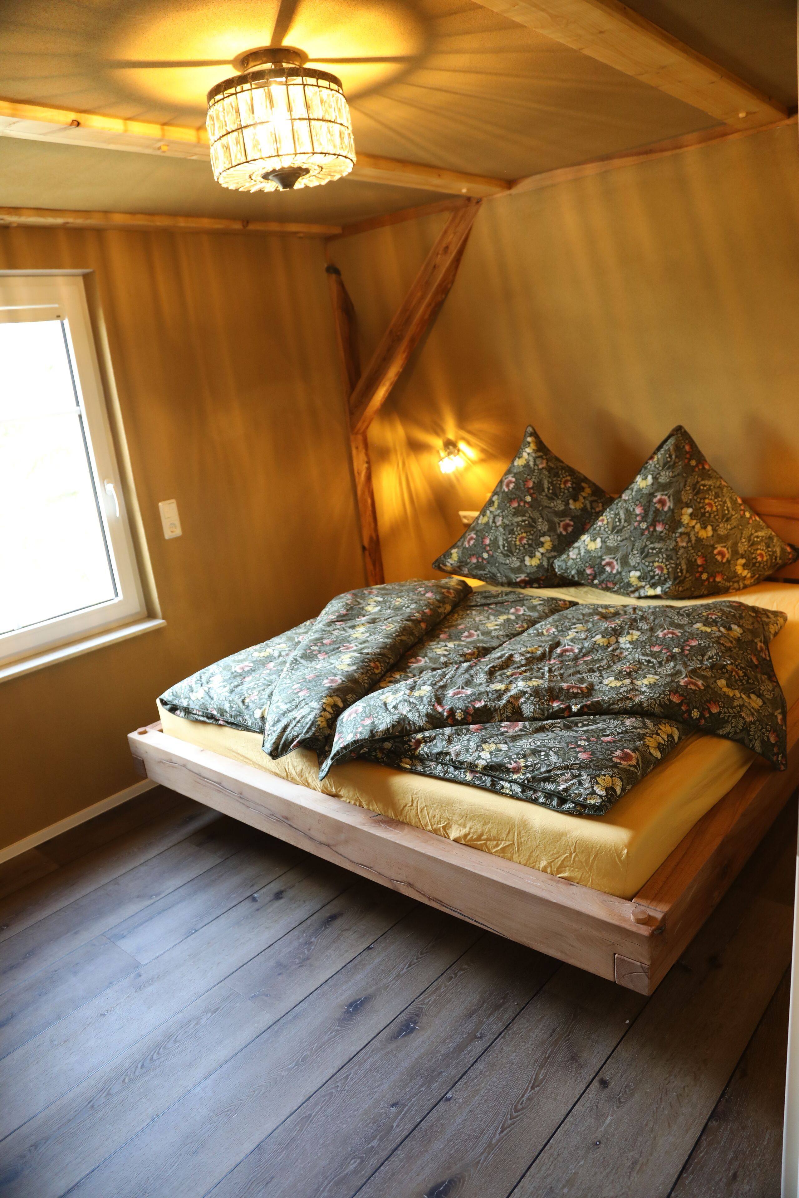 Una cama de madera con colcha y almohadas estampadas, una ventana con cortinas y una araña.