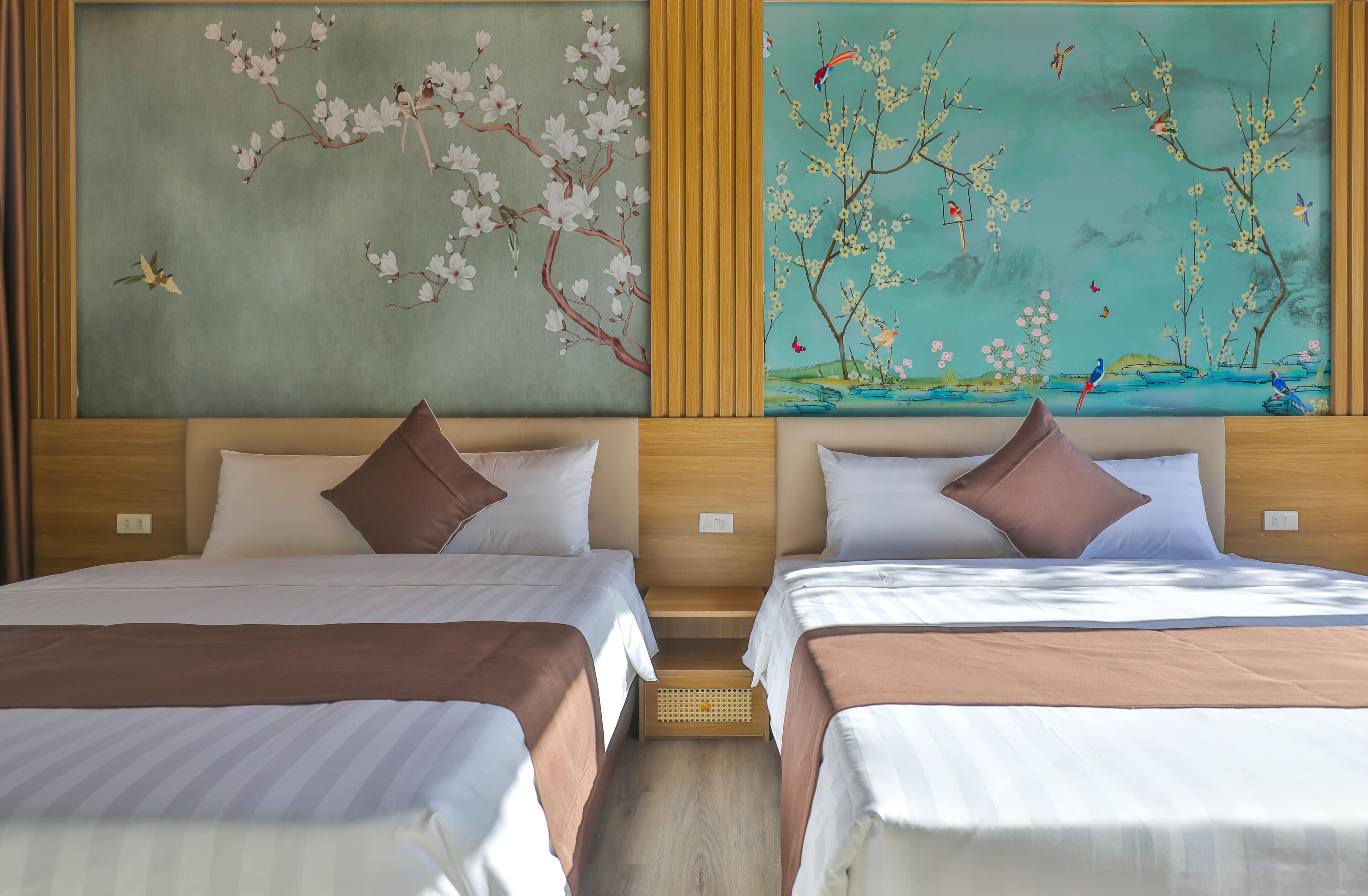 Hotelkamer met twee bedden, elk voorzien van een kussen en een bruine deken. De kamer heeft een houten hoofdbord en een groot schilderij met kersenbloesems en vogels.
